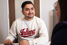 A male student in an IU sweatshirt talks with another person across a desk.